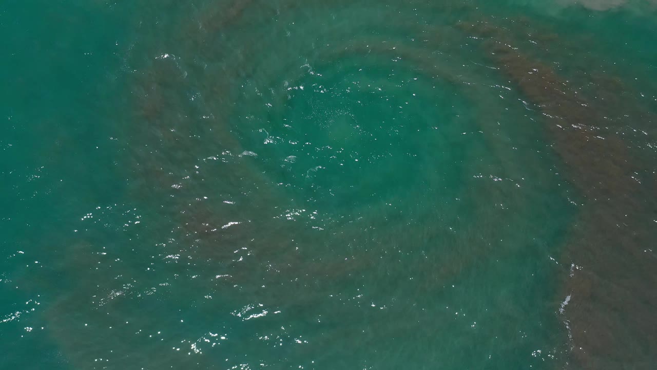 Aerial ocean frame over Los Cabos, Mexico showing teal water, scattered white highlights, and a subdued brown-pink eddy shape under the surface with no visible people.