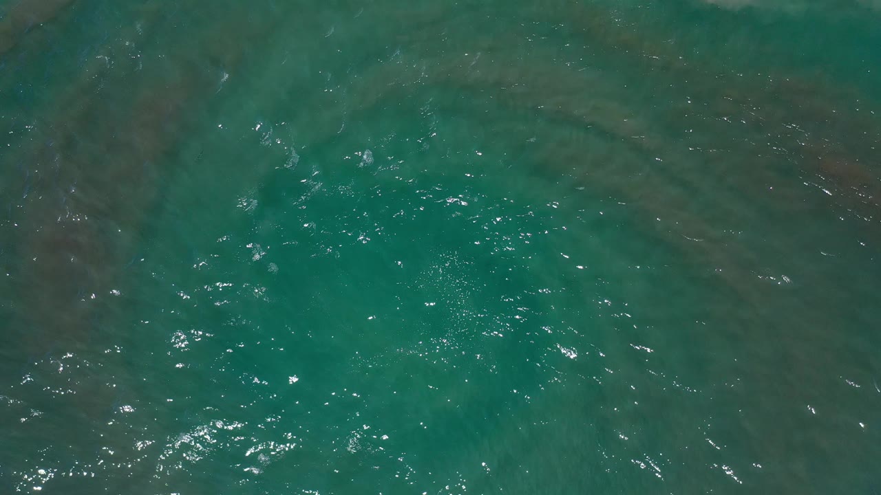 Overhead Los Cabos water frame showing turquoise-green surface movement, faint darker swirls below, bright specular flecks, and no visible people.