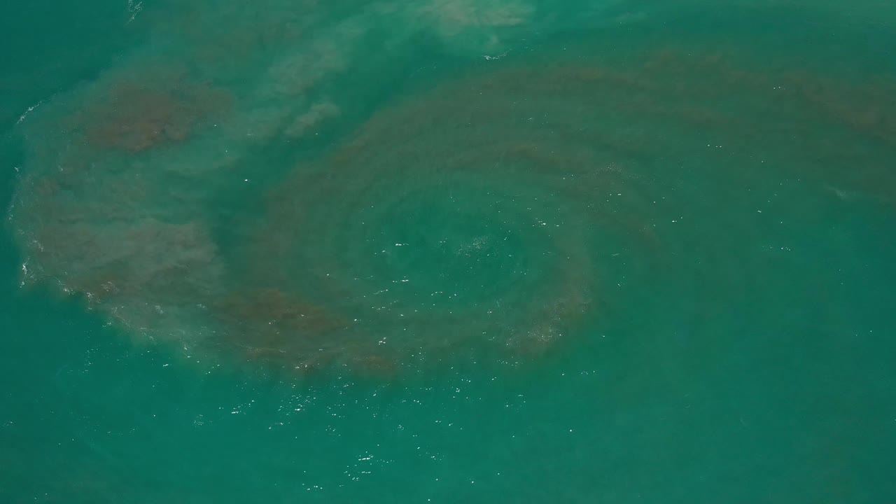 Aerial view over Los Cabos ocean water with the dark circular vortex shifted slightly left of center, pale surface glare, and no visible people.