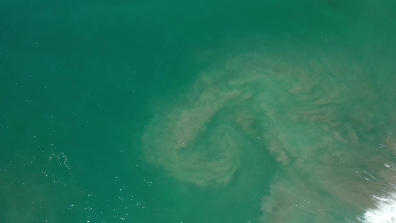 Aerial view near Los Cabos, Mexico, showing translucent green water, pale underwater formations, and a small white surf patch at the lower right with no visible people.