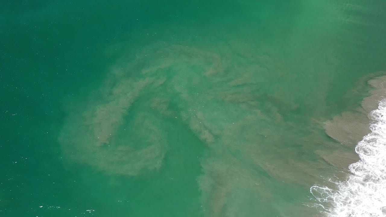Top-down view of green ocean water with pale sand patterns and surf at the right edge.