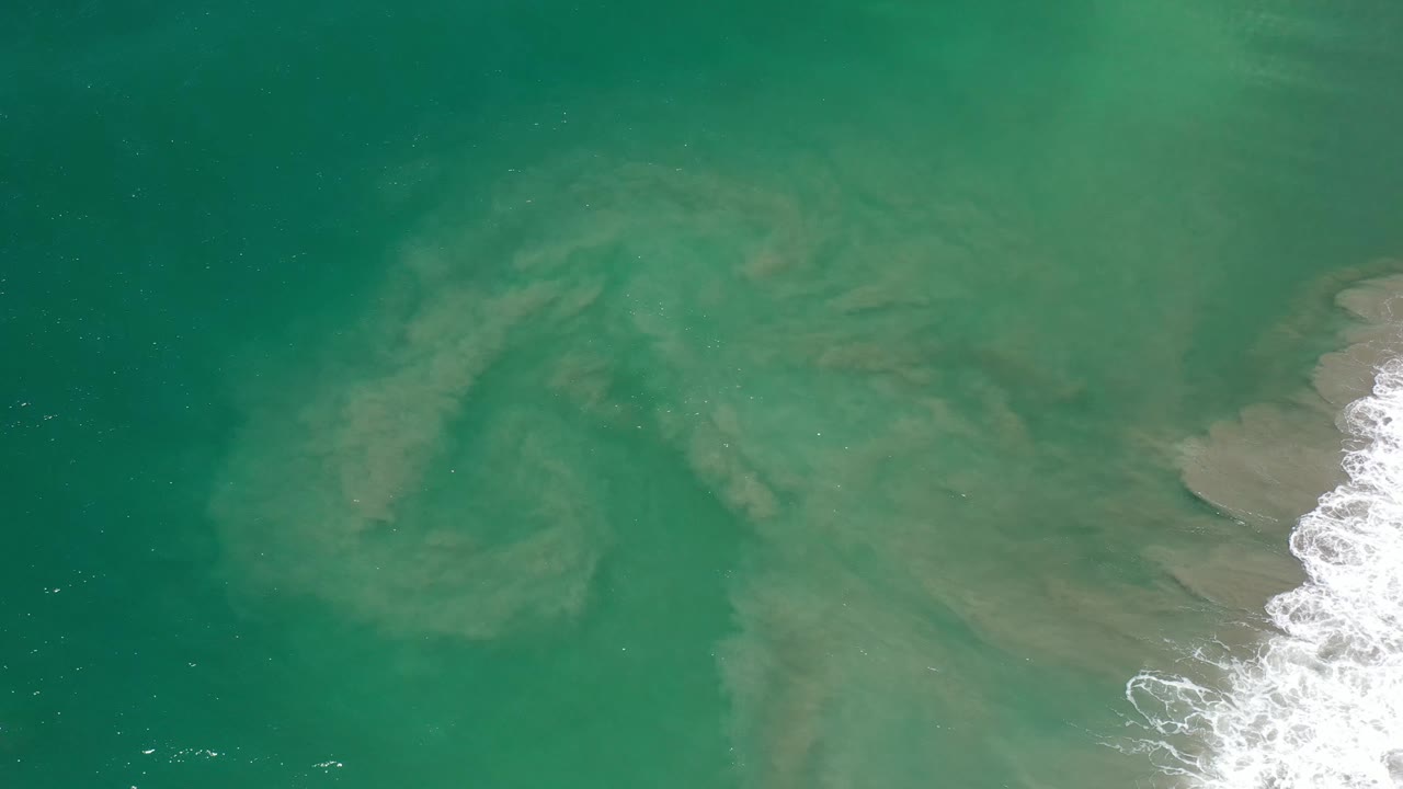 Bird's-eye frame from Los Cabos, Mexico, showing translucent green water over sand bars, a thin white foam band at the right margin, and no visible people.