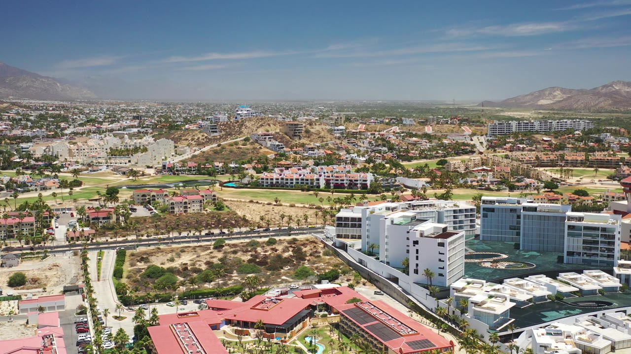 Wide aerial city view with modern buildings, red roofs, roads, and distant hills.