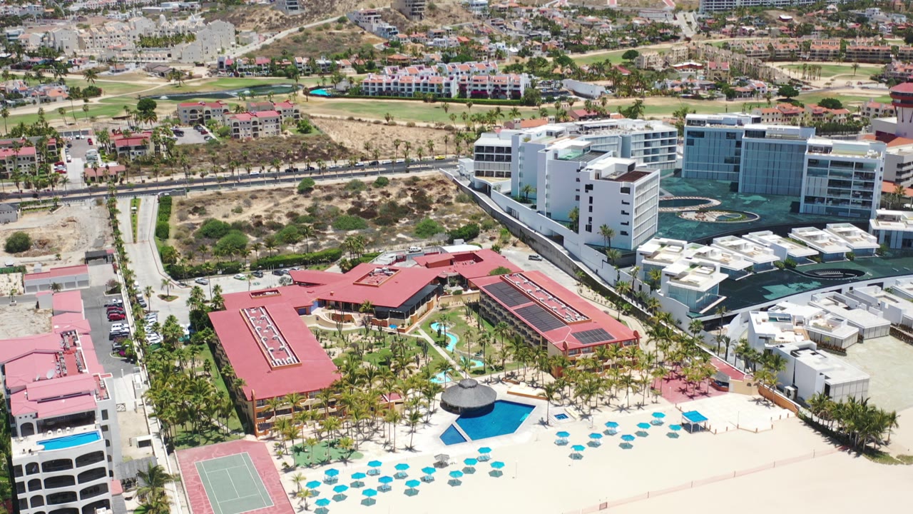 Aerial view in Los Cabos, Mexico, showing red-roofed beachfront buildings, palms, blue umbrellas, pale sand, roads, and modern white structures inland.
