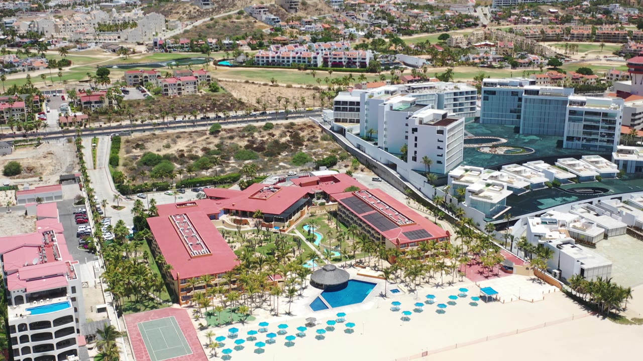 High aerial frame over Los Cabos coastal properties, with a pool and blue umbrellas near the sand, red roofs in the center, and roads and buildings beyond.