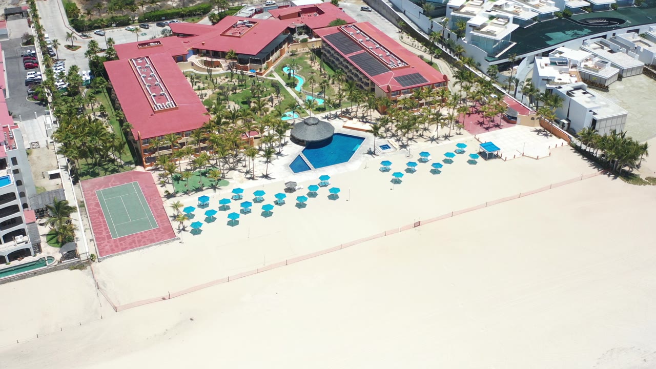 Aerial view of red-roof beach resort with pools, umbrellas, tennis court, and sand