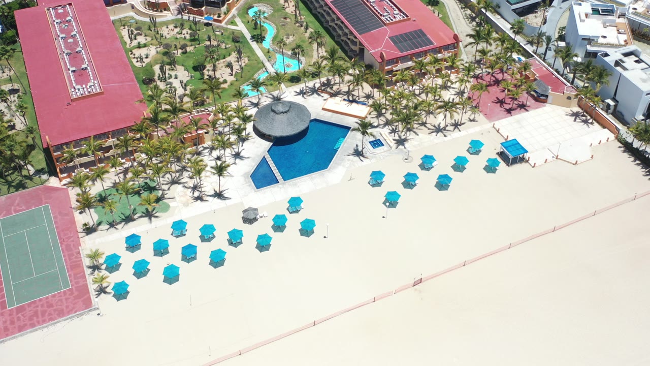 High aerial view of a Los Cabos resort beach with turquoise umbrellas on pale sand, a blue pool beside a round palapa roof, palm landscaping, red-roof buildings, tennis courts, and no visible people.
