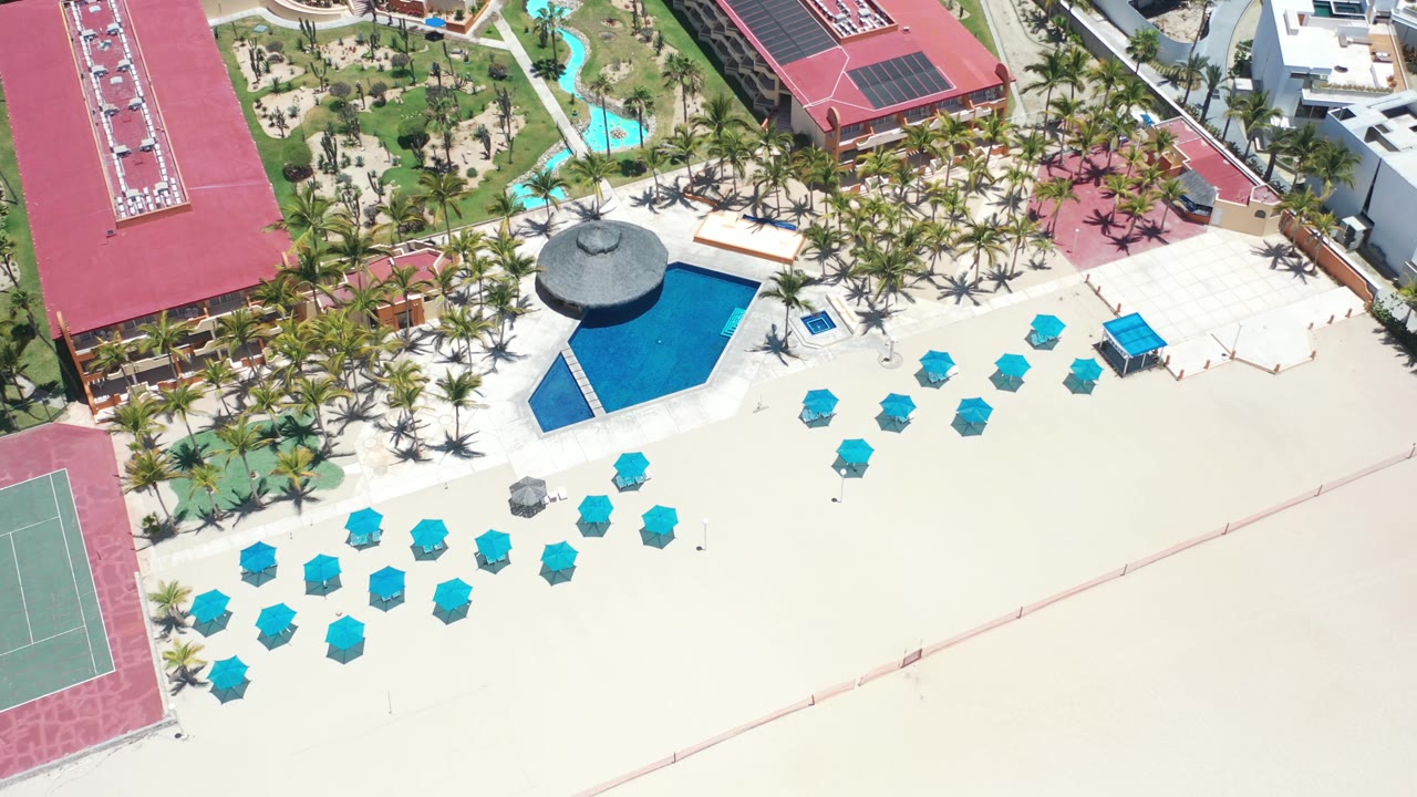 Wide overhead resort beachfront with blue umbrellas, pool, palms, and red roofs.