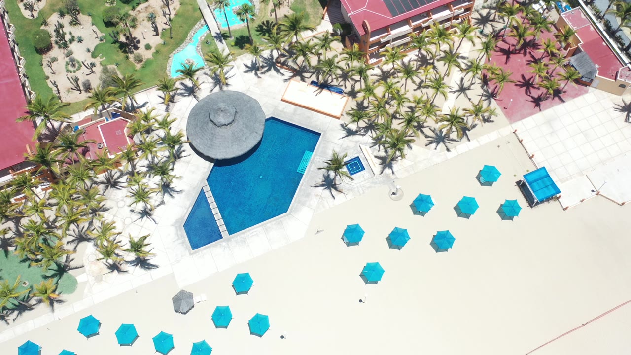 Straight-down aerial frame over a Los Cabos resort, showing a blue pool, round palapa roof, palm-lined grounds, teal beach umbrellas, pale sand, and no visible people.