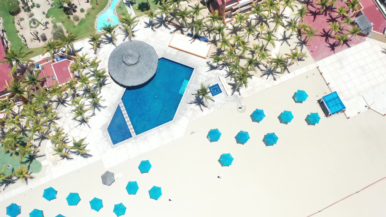 Top-down beach resort scene with pool, palms, umbrellas, and red-roof buildings.