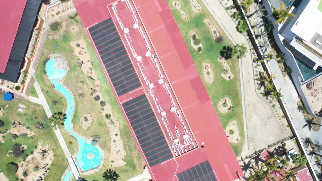 Top-down aerial frame in Los Cabos showing a red-roofed building beside a long marked court or roof deck, turquoise pools, lawns, palms, and no visible people.