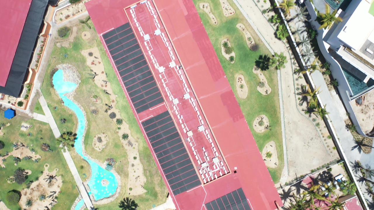 Aerial Los Cabos view centered on a red-roofed structure and dark rectangular court-like area, with pools and lawns to the side and no visible people.