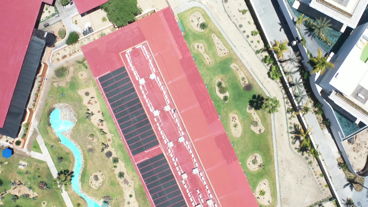 Vertical aerial view of a red roof, marked rectangular surface, pools, lawns, palms, and nearby modern buildings.