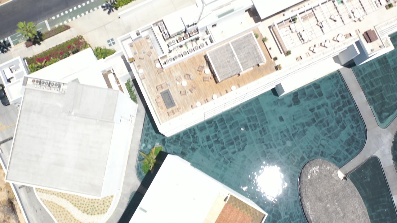 Top-down Los Cabos frame with white-roofed buildings around a blue-green pool, a paved road at the top edge, palm trees, hard sun glare on the water, and no clearly visible people.