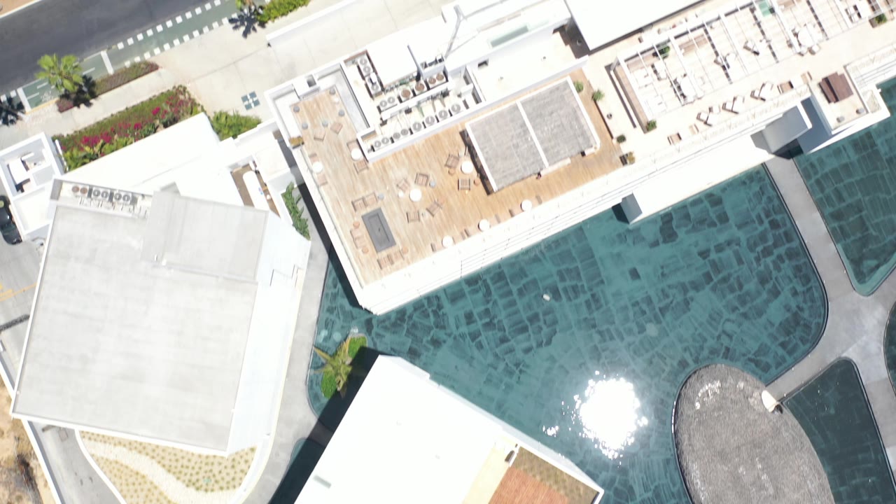 Top-down Los Cabos view showing the same pool shifted across frame, white building roofs, a curved pool edge, a dark road, sidewalk planting, and no clearly visible people.