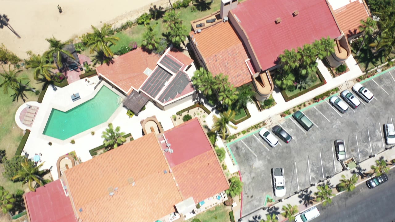 Overhead view of red-roofed buildings, pool, palms, parking lot, and sandy path.