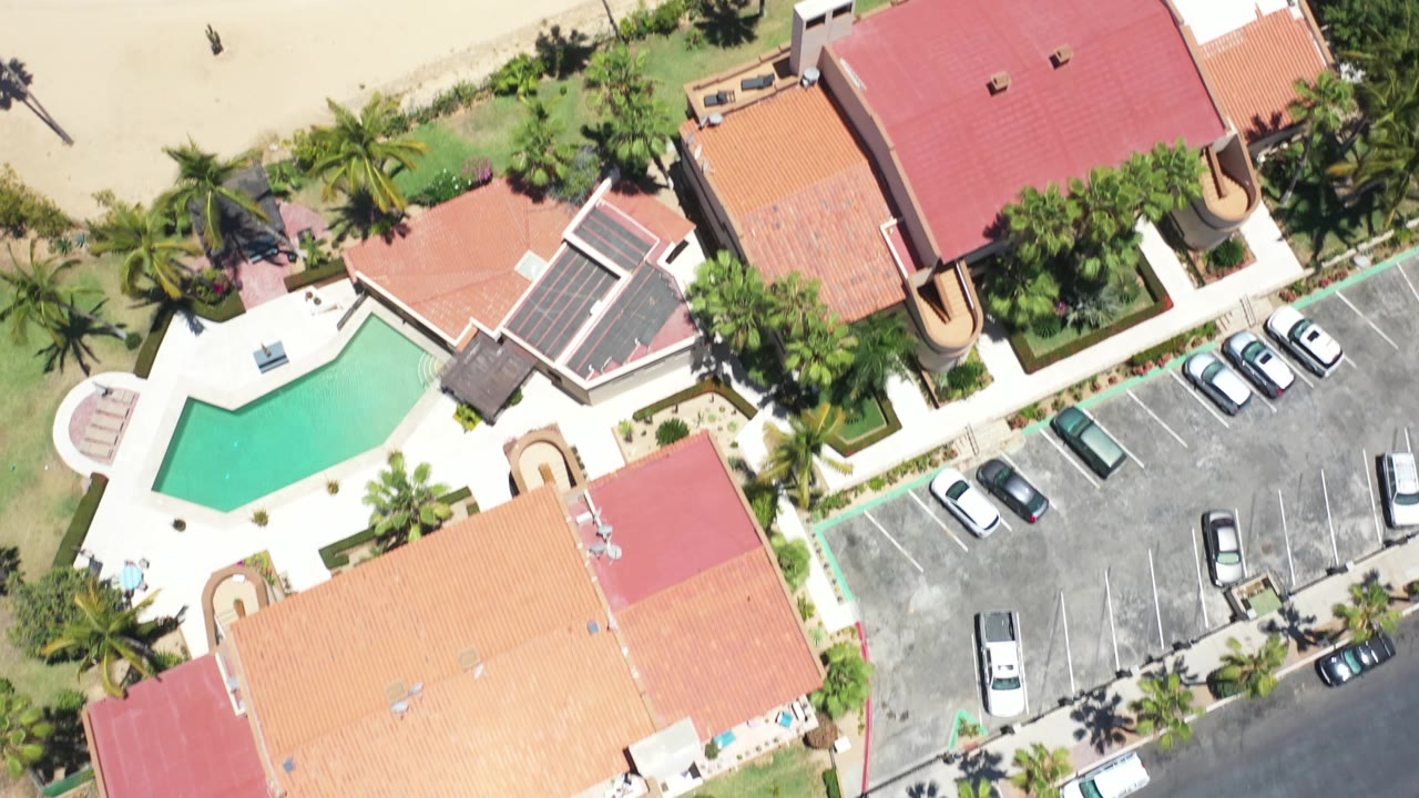 Los Cabos aerial view with the pool and red-roofed buildings shifted left of center, parked cars in a lot to the right, and no visible people.