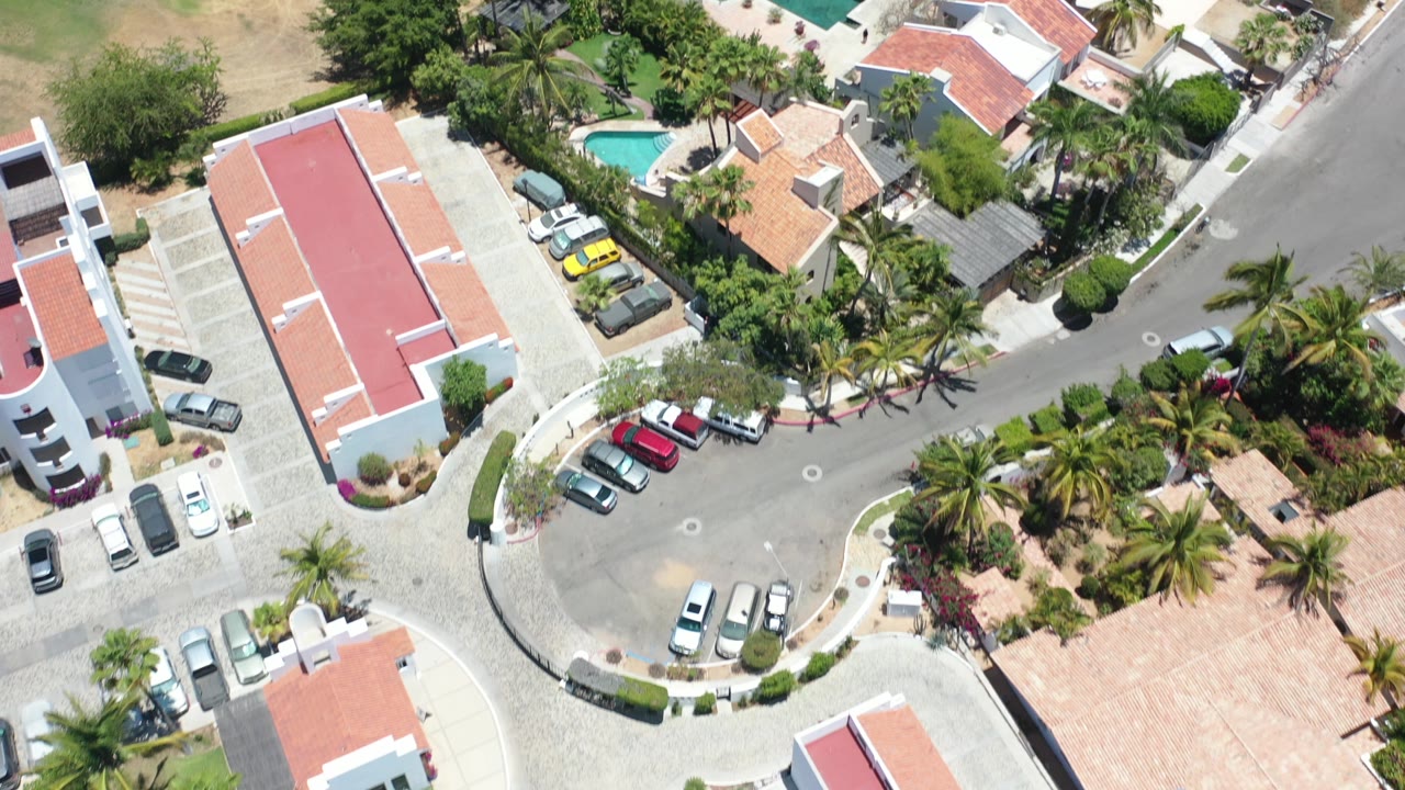 Overhead view of residential buildings, parked cars, streets, palms, and pools.