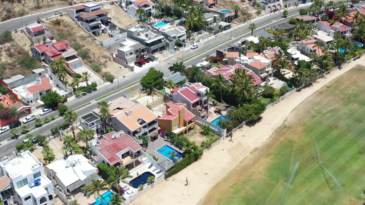 Aerial frame over Los Cabos, Mexico showing hillside houses, a paved road, palms, pools, dry slopes, and a green landscaped strip with no visible people.