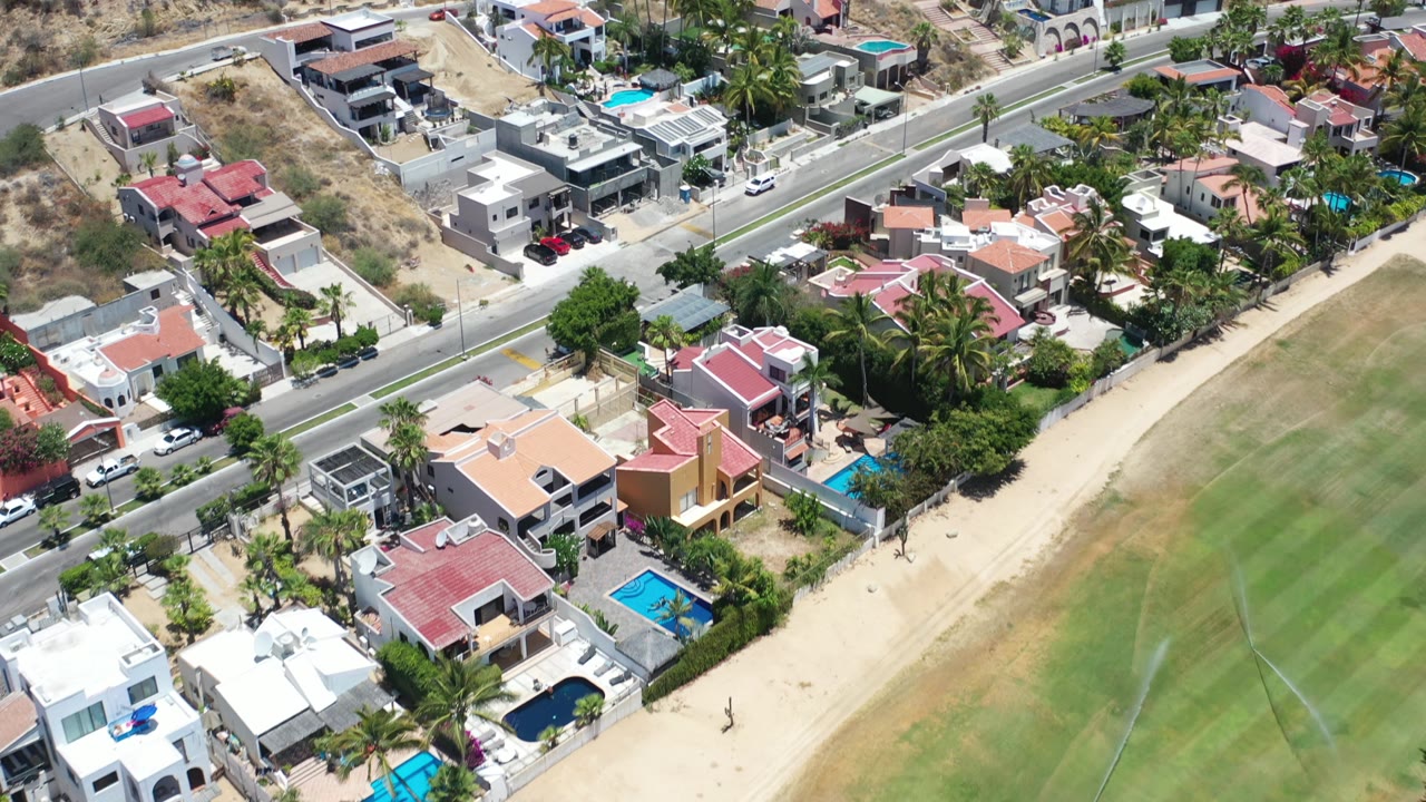 Aerial view in Los Cabos, Mexico with red and white rooftops, blue pools, palm trees, parked cars, a road, and dry hillside terrain with no visible people.