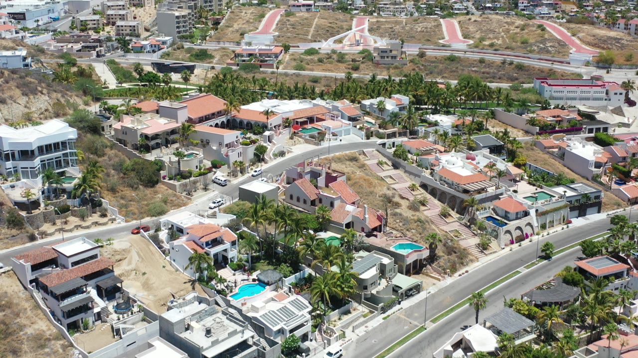 Aerial view over villas, curved roads, pools, palms, dry hills, and city buildings.