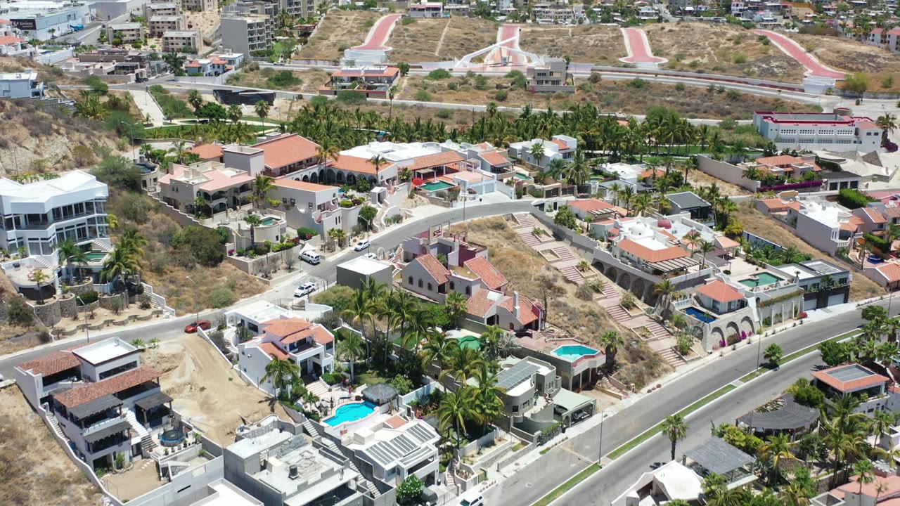 Aerial Los Cabos frame with foreground villas and pools, a curving road, palm trees, dry hillside landscaping, city buildings in the distance, and no visible people.