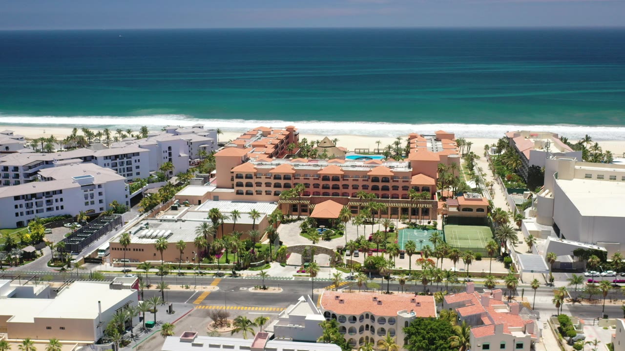 Wide aerial view in Los Cabos, Mexico, with turquoise ocean and surf behind beachside hotel buildings, palm-lined streets, rooftops, and no close visible people.