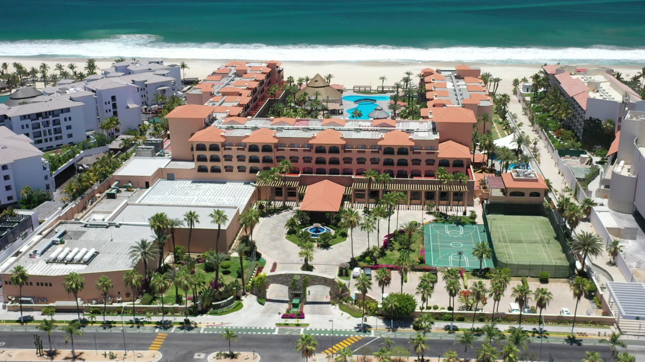 Opening aerial frame over Los Cabos, Mexico showing terracotta hotel roofs, a blue pool, sports courts, palm landscaping, beachfront road, surf, and no individually recognizable people.