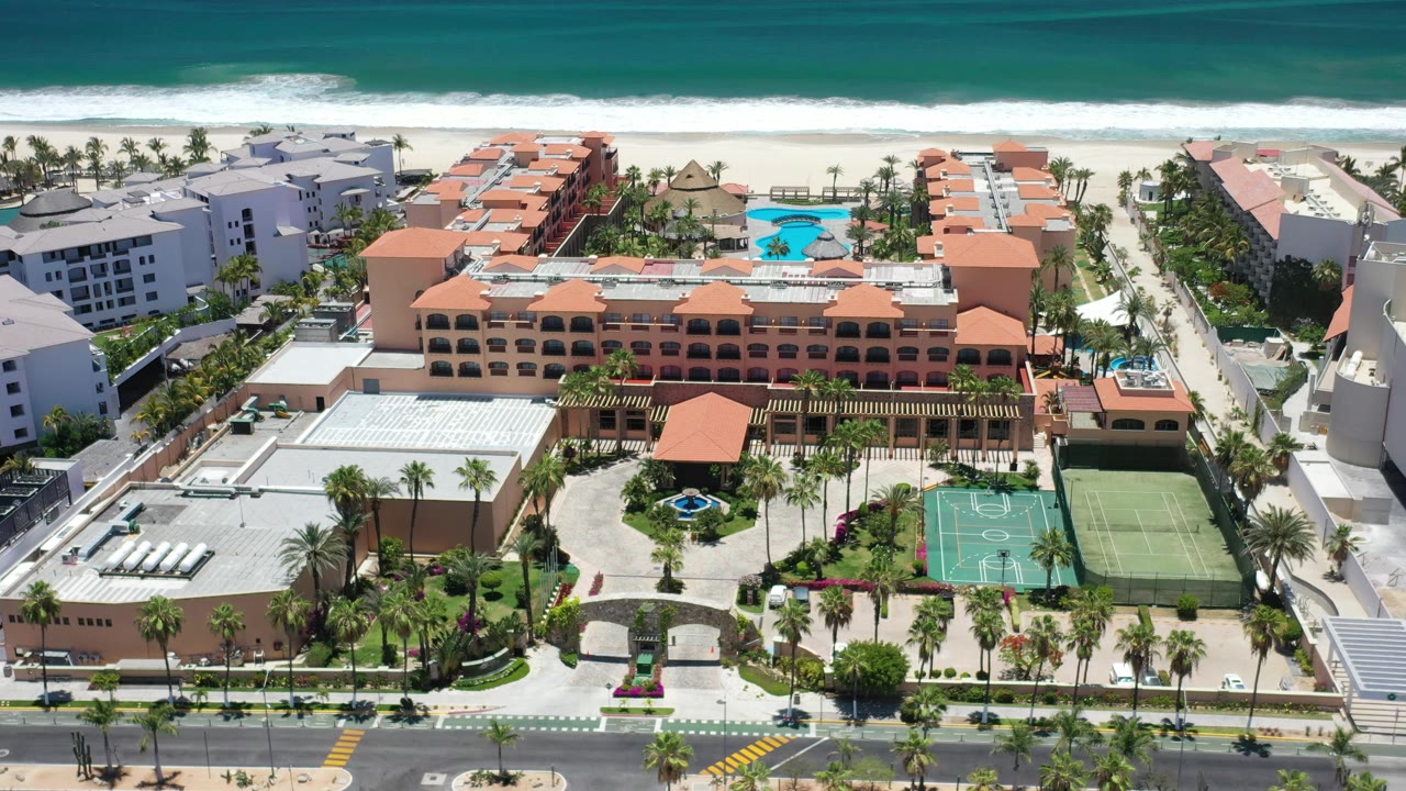 Second Los Cabos aerial frame with the blue pool slightly farther right, orange-roof hotel blocks centered, sports courts in the lower right, beach and white surf across the top.