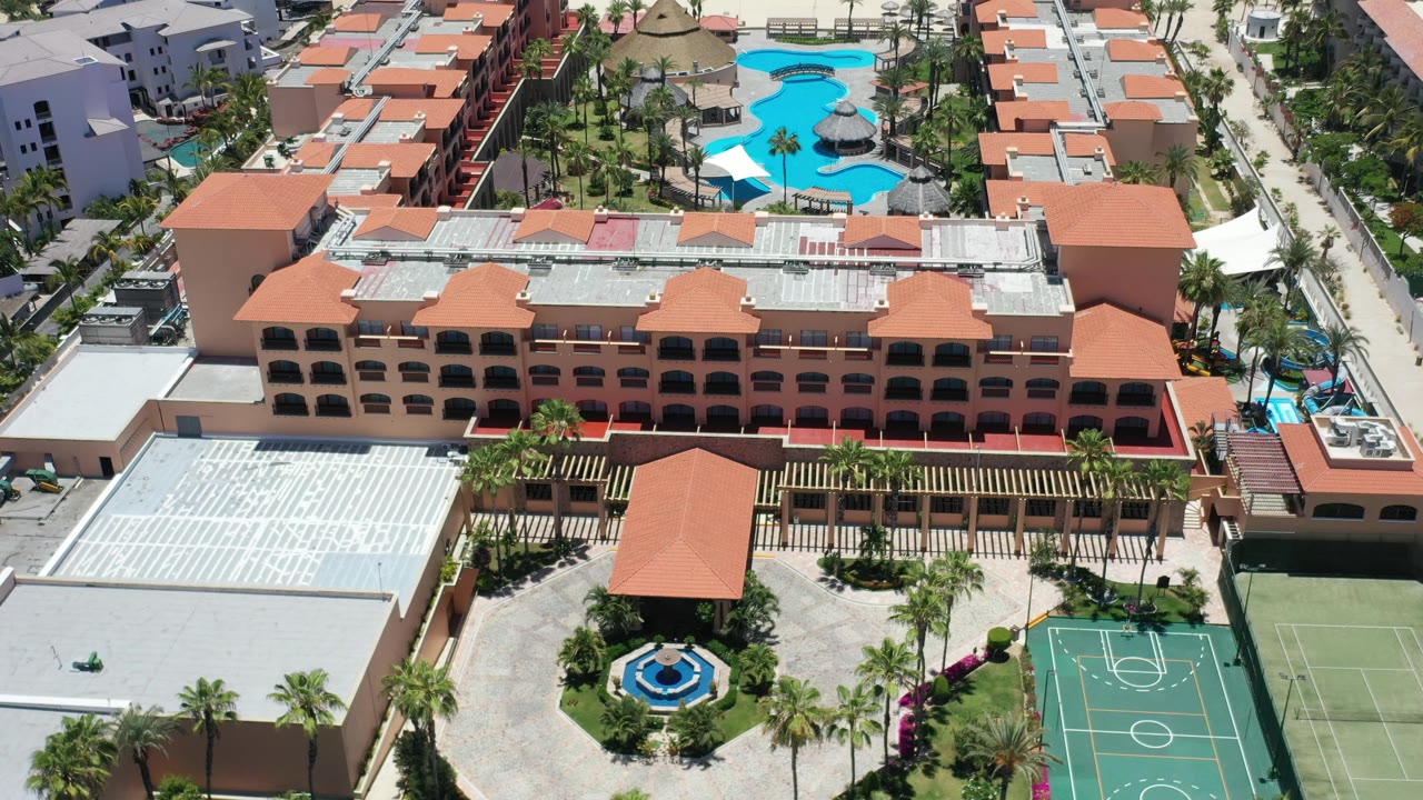 This Los Cabos resort frame keeps the main terracotta building centered, with the bright pool area above it, palms along the walks, a basketball court at lower right, and no visible people.