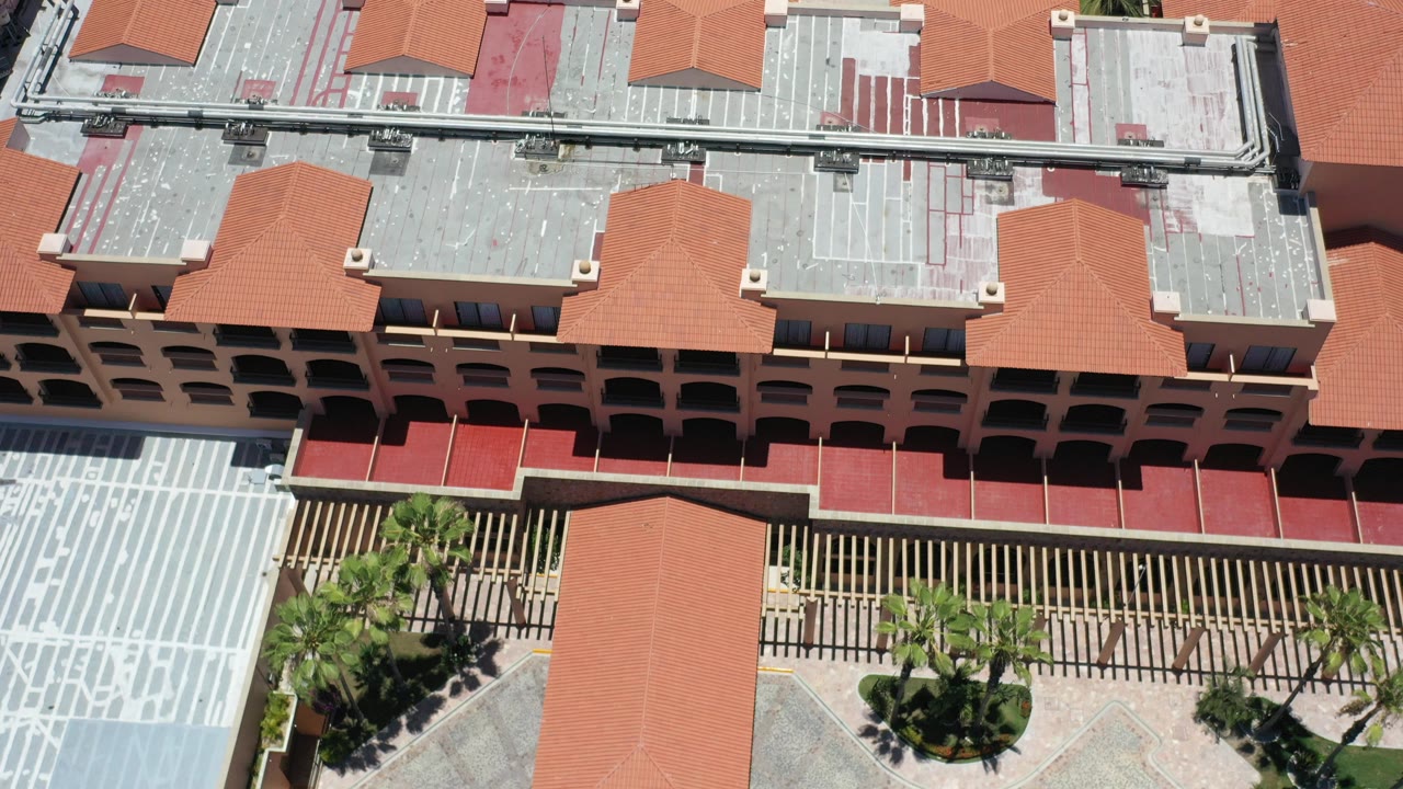 High-angle aerial frame over a Los Cabos resort roofline, with red-tile roofs, flat service rooftops, palm landscaping, and no visible people.