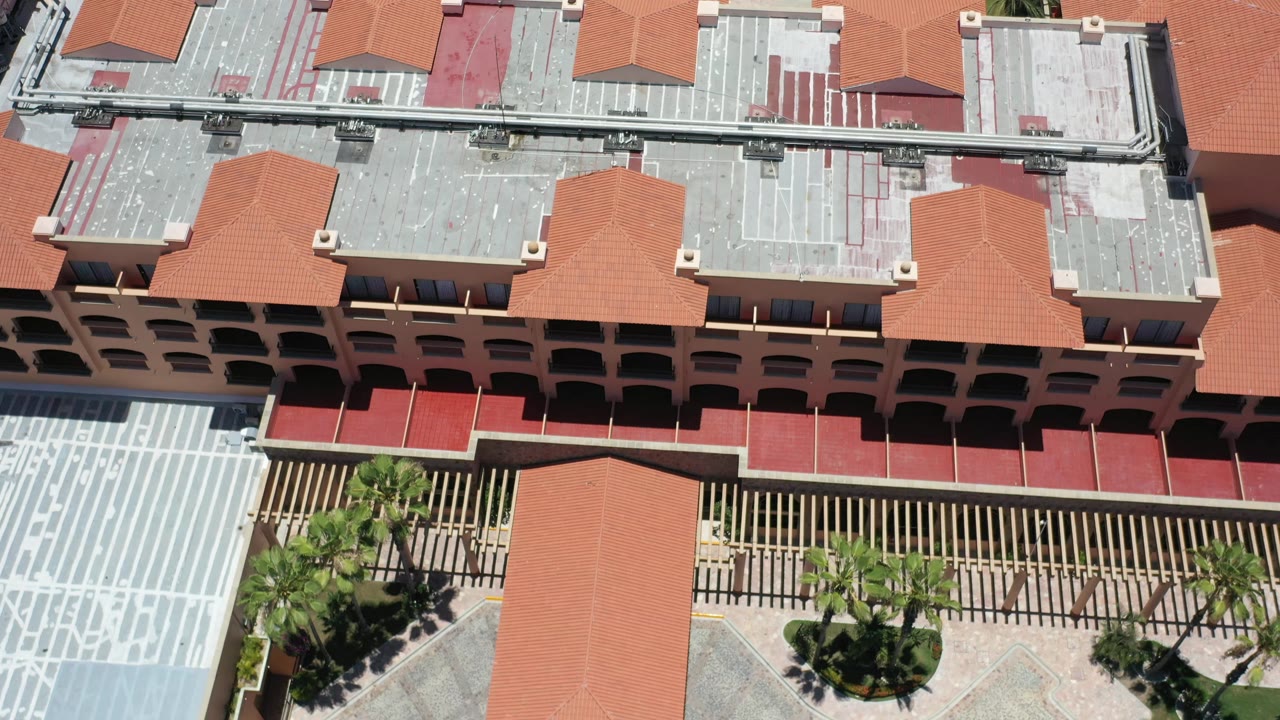 A second high-angle view of the Los Cabos hotel roofs shows red awnings, balcony rows, palm trees, and patterned courtyard paving with no visible people.