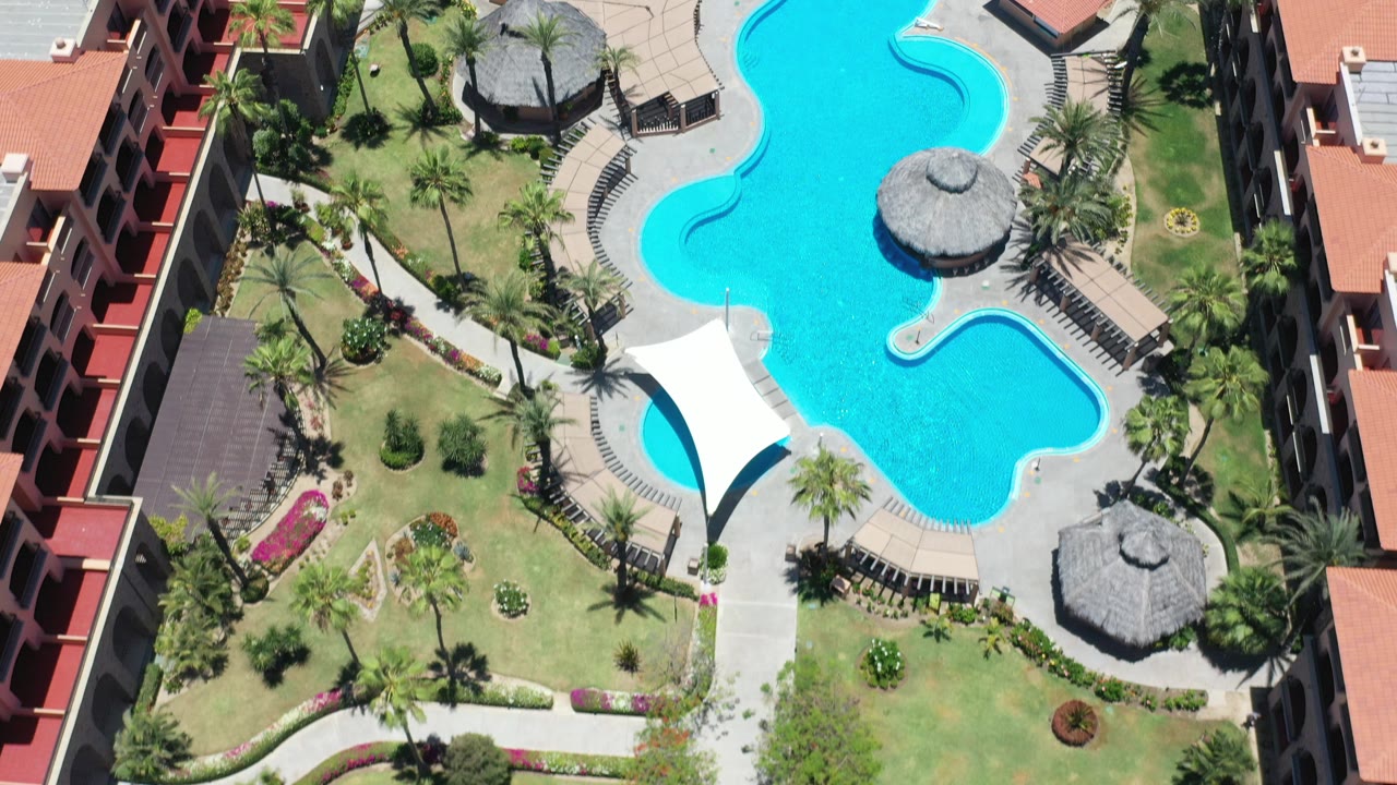 Overhead view in Los Cabos, Mexico, showing a turquoise resort pool between red-roofed buildings, palms, paths, and thatched shade structures, with no visible people.