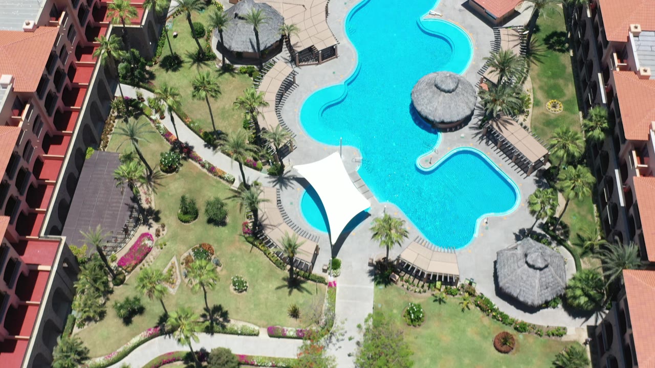 Aerial frame from Los Cabos, Mexico, with the curved pool centered beside landscaped grass, palm trees, red-roofed buildings, and a white canopy, with no visible people.