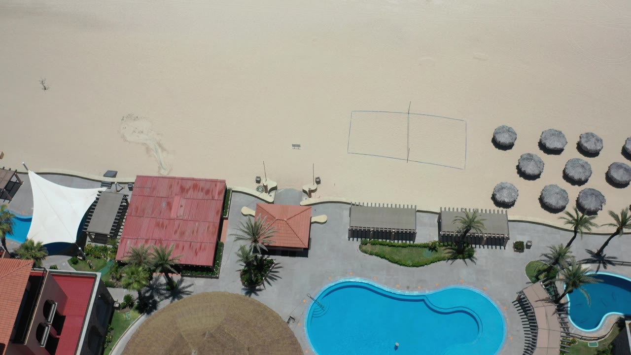 Overhead beach club area with pools, red roofs, white canopy, umbrellas, and sand.