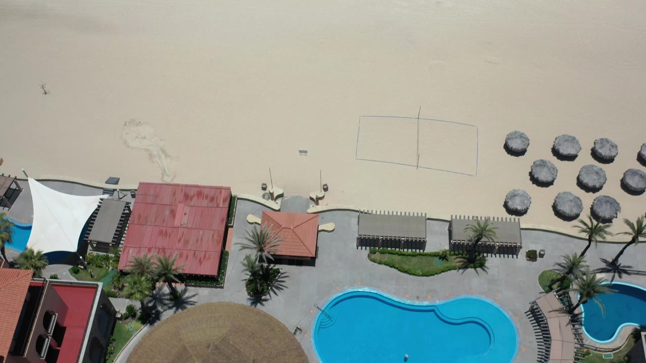 Aerial frame in Los Cabos, Mexico with blue pools at the lower edge, red roof buildings, white canopy fabric, clustered beach umbrellas, open sand, and no visible people.