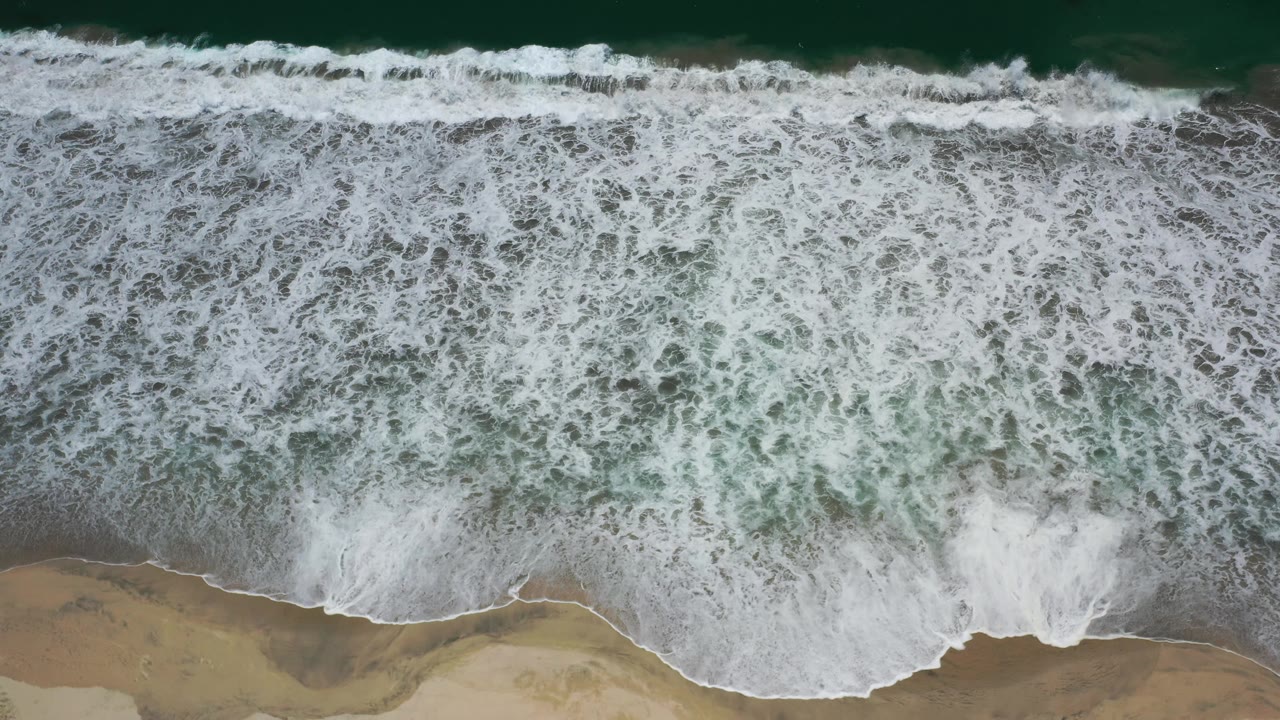Aerial view of a broad white wave line pushing toward the sandy Los Cabos shore, with darker green water beyond and no visible people.