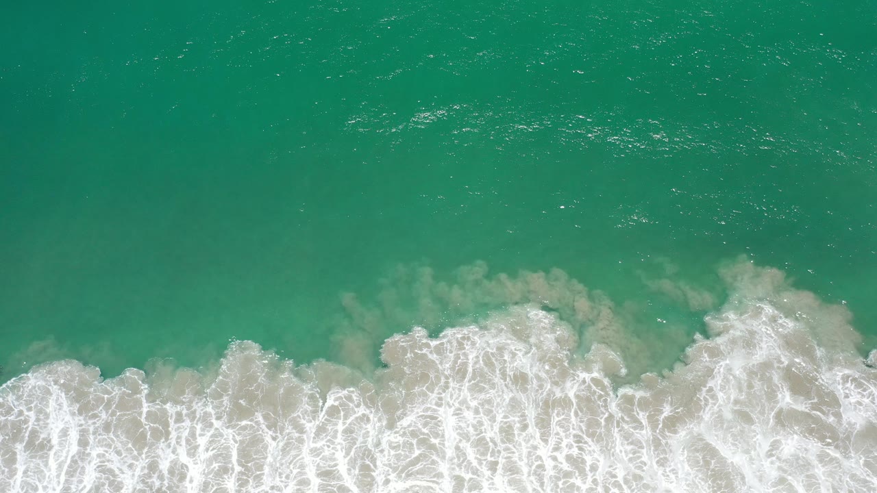 Overhead view in Los Cabos, Mexico of turquoise green water meeting a wide band of white surf over pale sand, with no visible people.