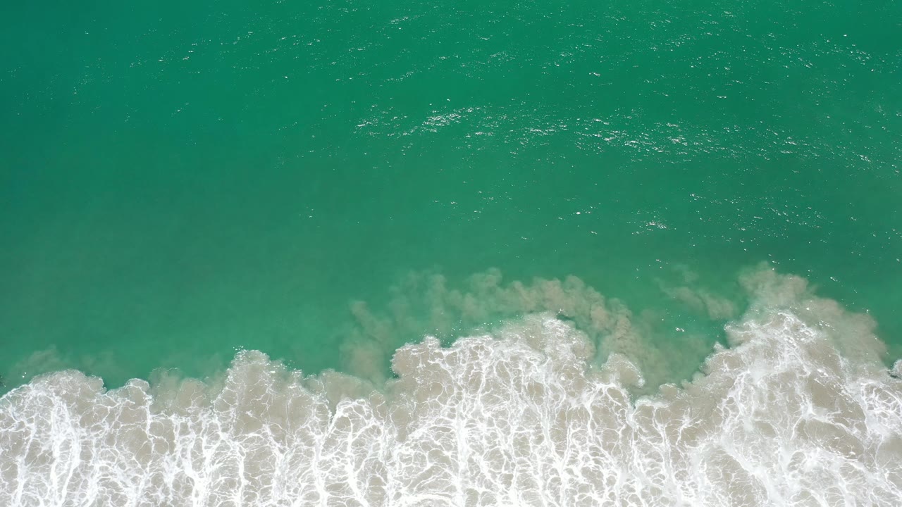 Top-down Los Cabos shoreline frame with white wave foam pushed farther across the pale sand and green water above, with no visible people.