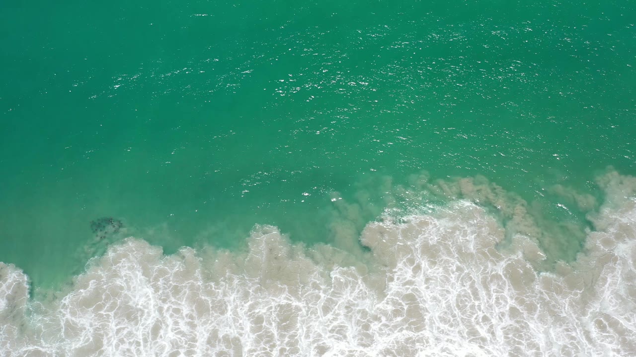 A band of white foam pushes farther across the lower frame beneath green ocean water in Los Cabos, Mexico, with no visible people.