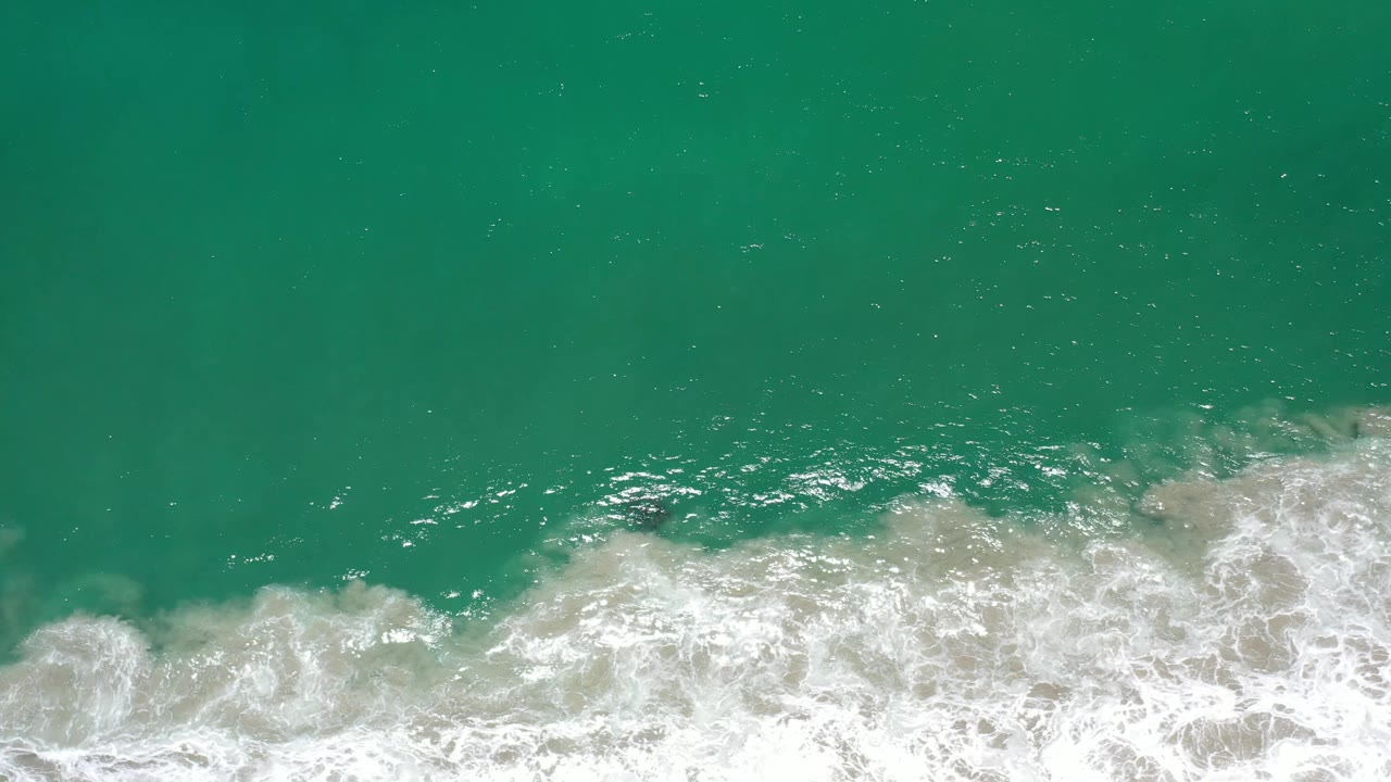 Aerial frame over Los Cabos green water, with sun flecks across the surface and a thicker line of white foam entering from the lower edge, no visible people.