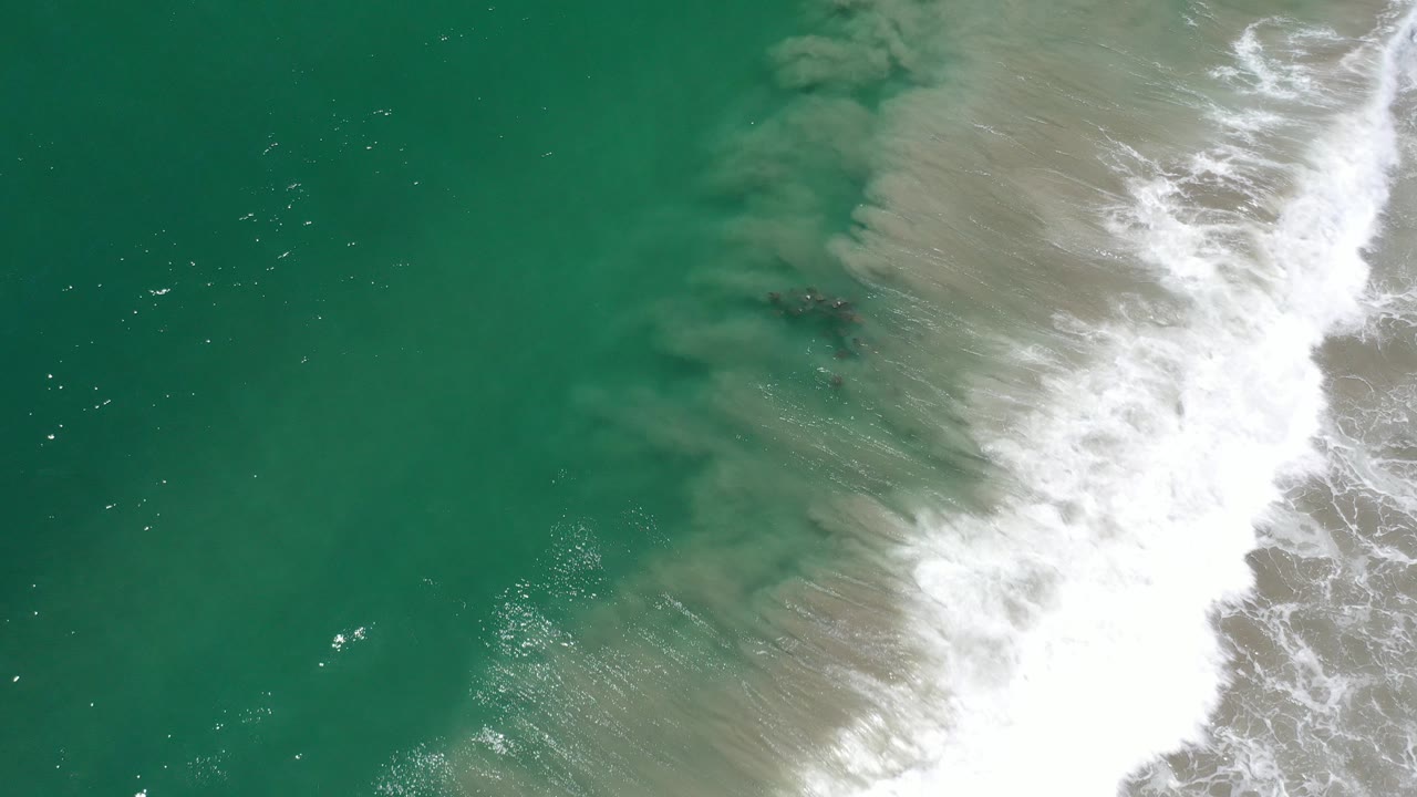 Overhead view in Los Cabos, Mexico of turquoise water on the left and a broad white surf break pushing across sand-tinted shallows on the right, with no visible people.