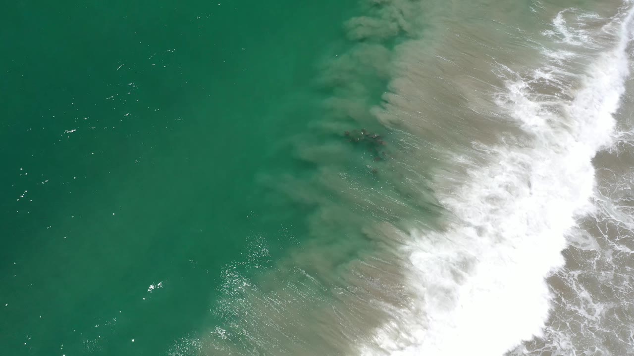 Aerial frame from Los Cabos, Mexico showing green-blue ocean water, tan suspended sand near the break, and white foam clustered along the right side, with no visible people.