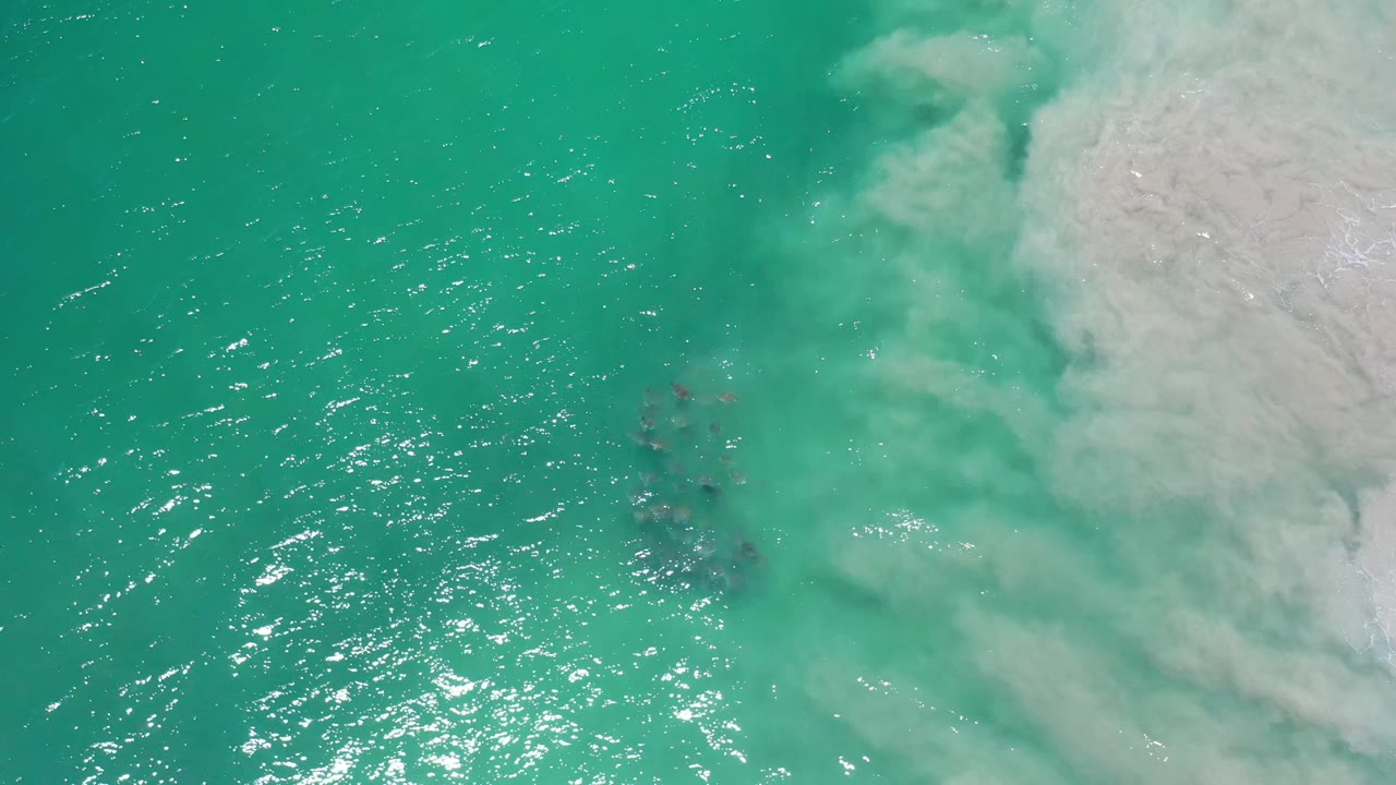 Overhead Los Cabos ocean frame with bright surface reflections across the left side, a soft foam bank on the right, faint submerged rocks near the middle, and no visible people.
