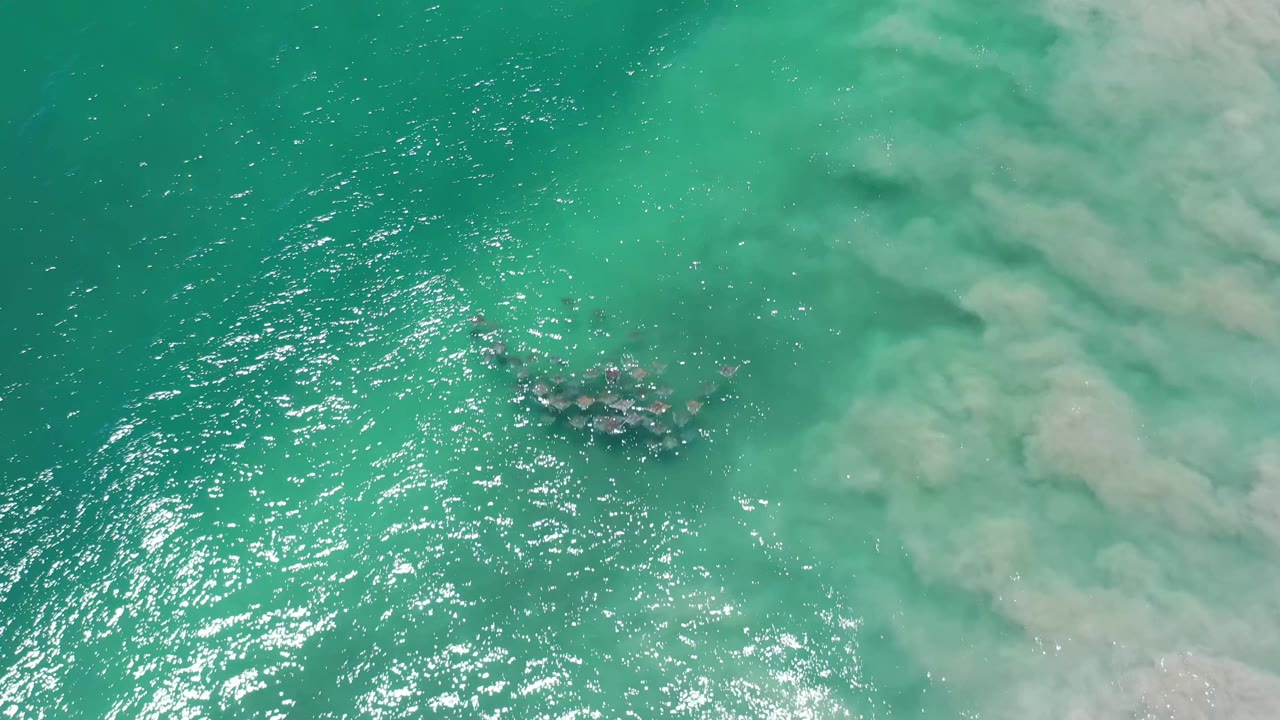 Aerial Los Cabos frame with the dark cluster slightly clearer against turquoise water, bright ripples on the left, and pale cloudy patterns on the right, with no visible people.