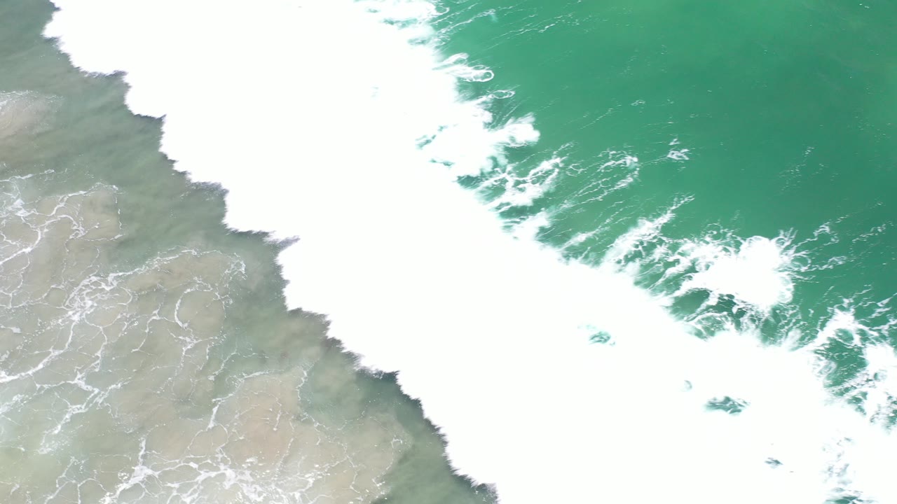 Overhead view in Los Cabos, Mexico of green water and dense white surf pushing diagonally over pale wet sand, with no visible people.