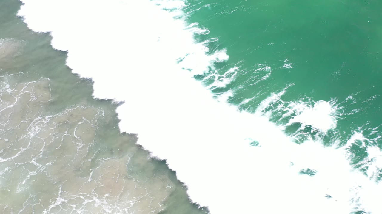Overhead Los Cabos coastal frame with a broad band of white foam over green water and a strip of wet sand at the lower left, with no visible people.