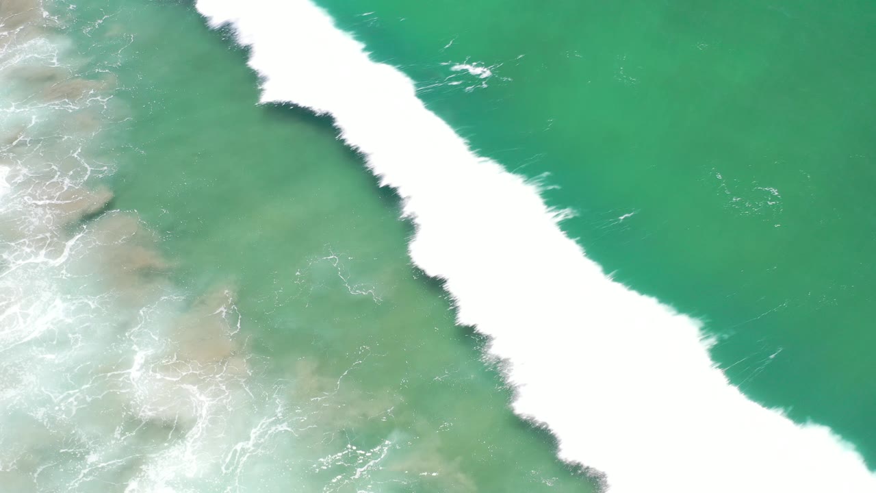 A bright white wave line runs diagonally through green Los Cabos water, with pale rocky shallows on one side and no visible people.