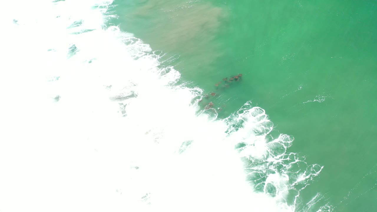 Overhead view of turquoise water in Los Cabos, Mexico, with white surf entering from the left, dark submerged reef or rocks near center, and no visible people.
