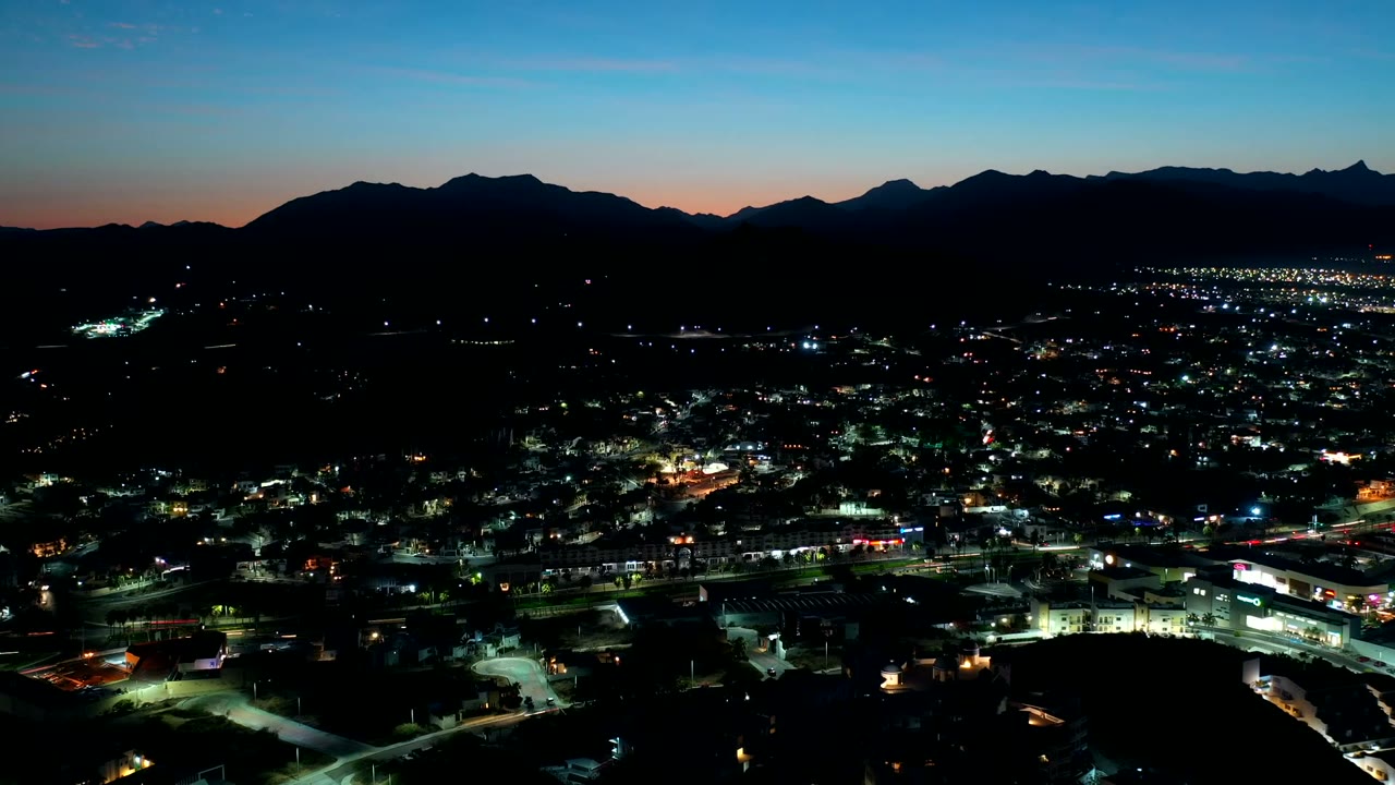 Aerial view over Los Cabos, Mexico at night, with scattered town lights below a black mountain silhouette and blue dusk sky, no visible people.